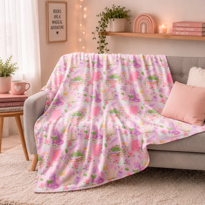Manta literaria con diseño de dragones lectores, libros y flores en tonos rosados, confeccionada en felpa con reverso sherpa blanco.