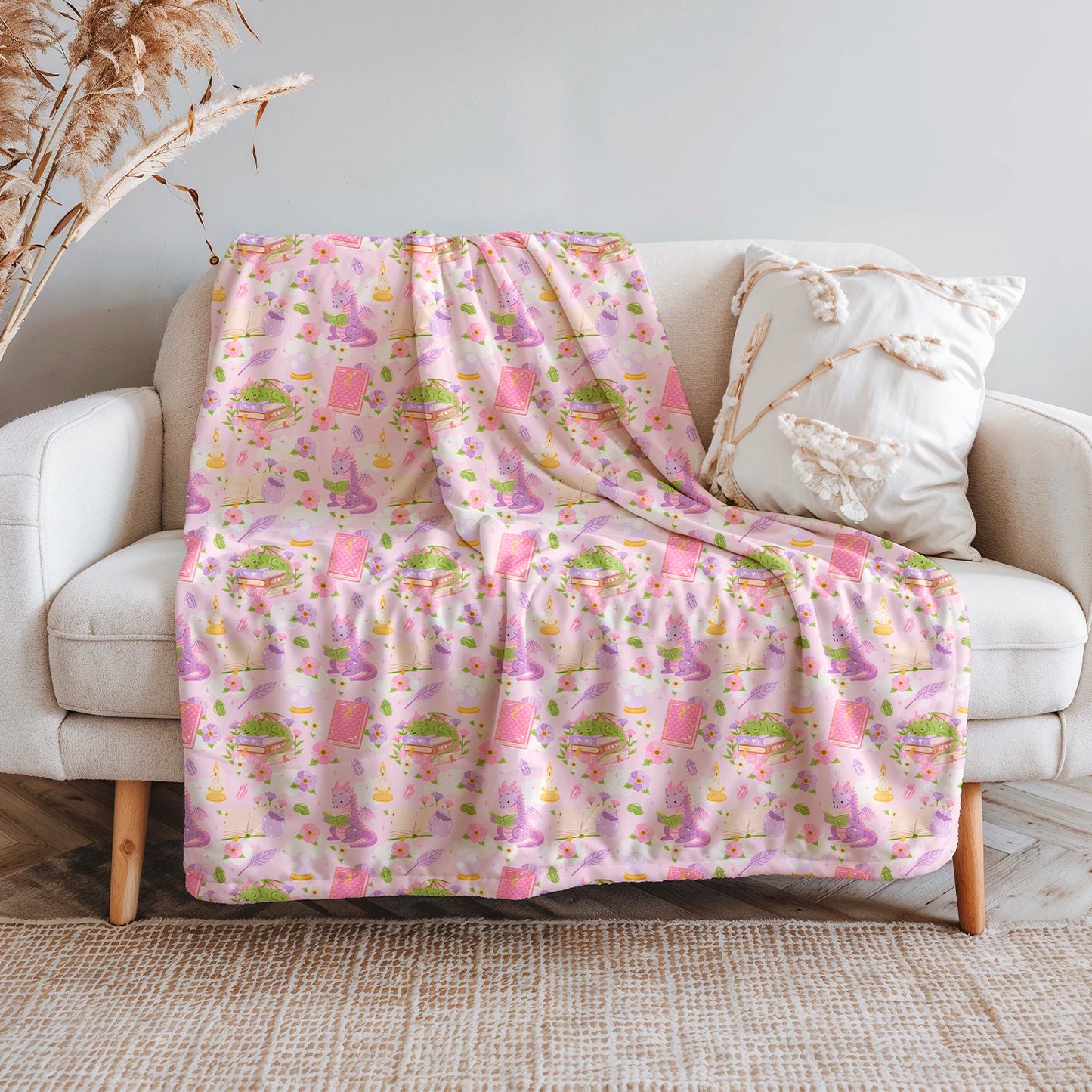 Manta literaria con diseño de dragones lectores, libros y flores en tonos rosados, confeccionada en felpa con reverso sherpa blanco.