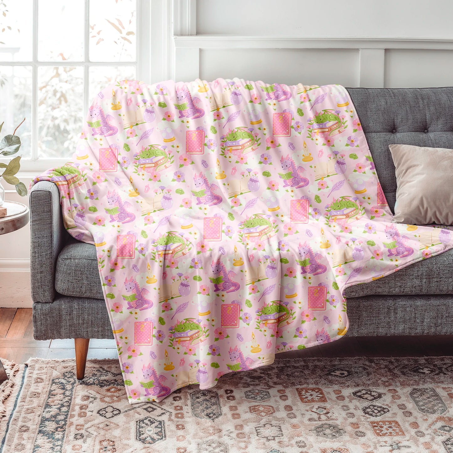 Manta literaria con diseño de dragones lectores, libros y flores en tonos rosados, confeccionada en felpa con reverso sherpa blanco.