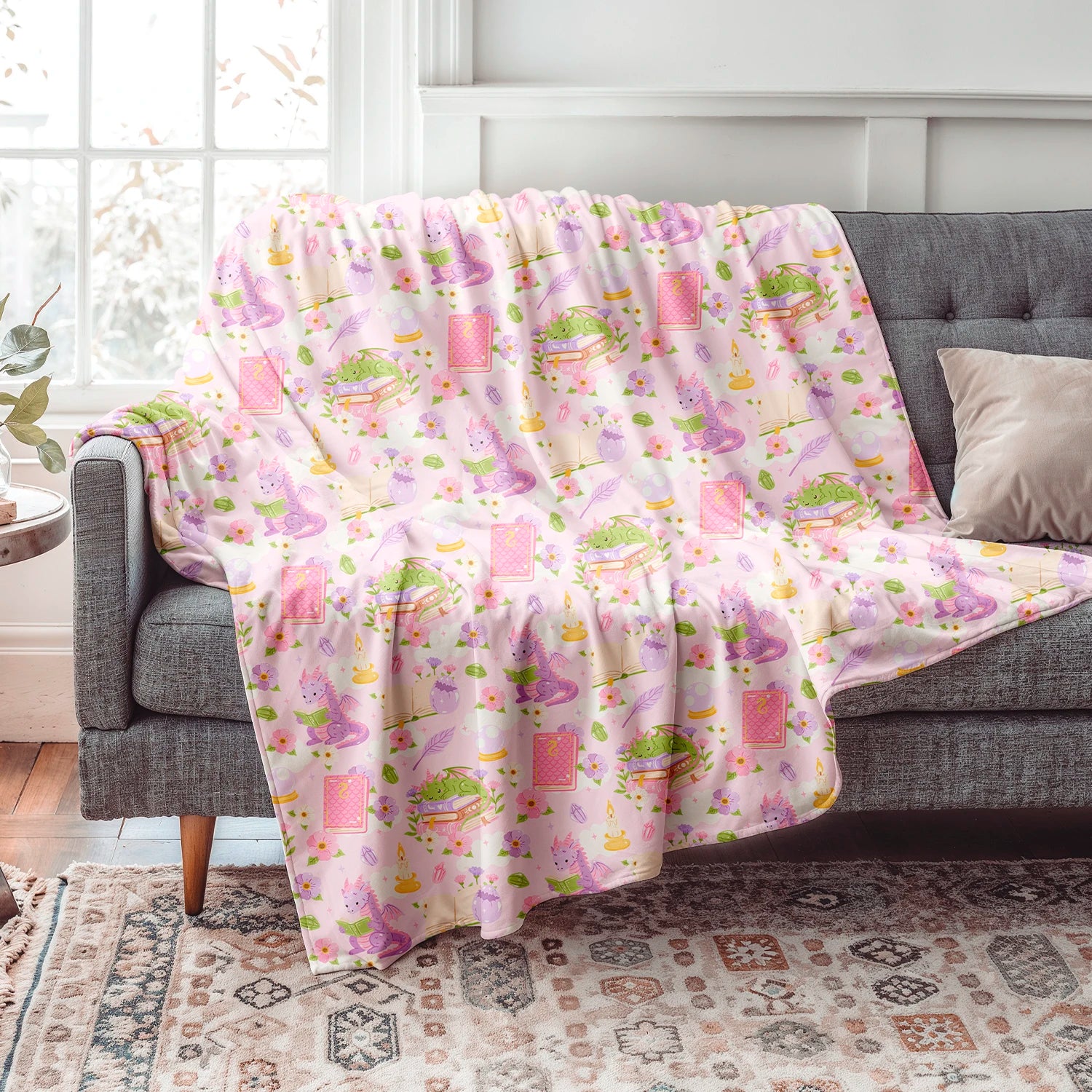 Manta literaria con diseño de dragones lectores, libros y flores en tonos rosados, confeccionada en felpa con reverso sherpa blanco.