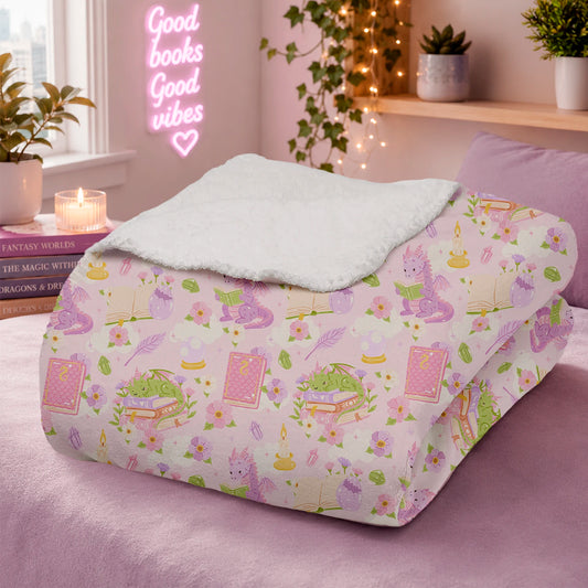 Manta literaria con diseño de dragones lectores, libros y flores en tonos rosados, confeccionada en felpa con reverso sherpa blanco.